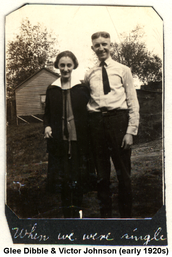 Black and white photo of Glee Dibble and Victor Johnson standing together on a sloping hillside and smiling; Glee, with bobbed dark hair wears a blouse with a wide white collar and a dark skirt; Victor, hair parted in the middle, wears a white long-sleeve shirt and short dark tie. Behind them is a frame building and trees. The photo is affixed to a black album page, on which is written in white ink 'When we were single'.